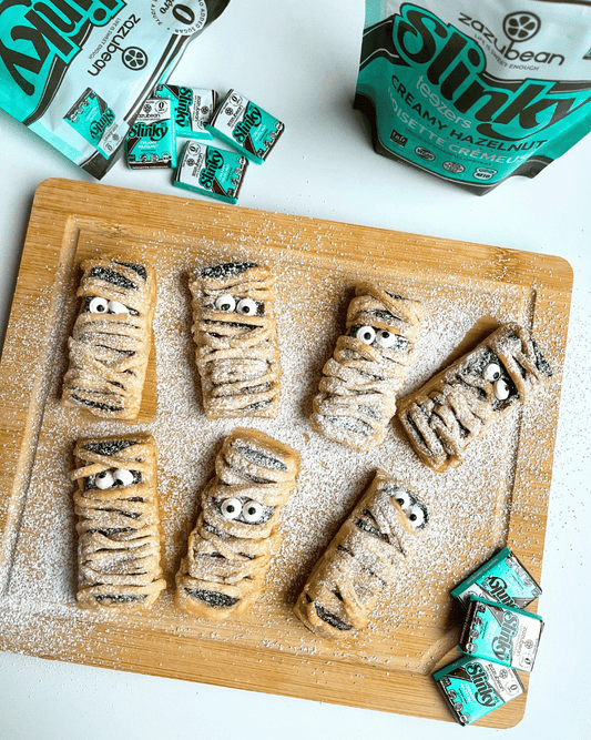 puff pastry mummies on a wooden cutting board with zazubean chocolates laying around the counter