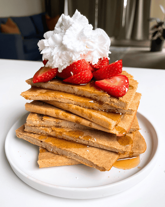 sheet pan vegan protein pancakes piled on a white plate with strawberries, maple syrup, and whipping cream
