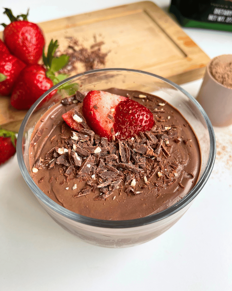 Silken Tofu Chocolate Pudding In a Glass container topped with a strawberries and chocolate pieces with some strawberries on a wooden cutting board in the background