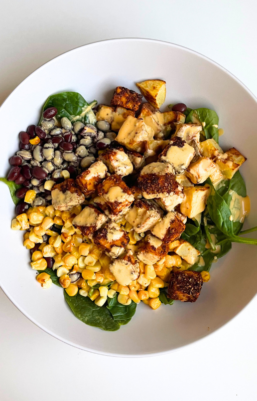 Spinach salad topped with black beans, yellow corn, baked tofu and chilli oil in a white plate on a white background