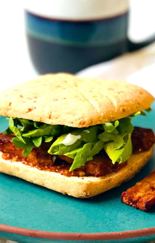 A tempeh sandwich on top of a green plate with a blurry mug in the background