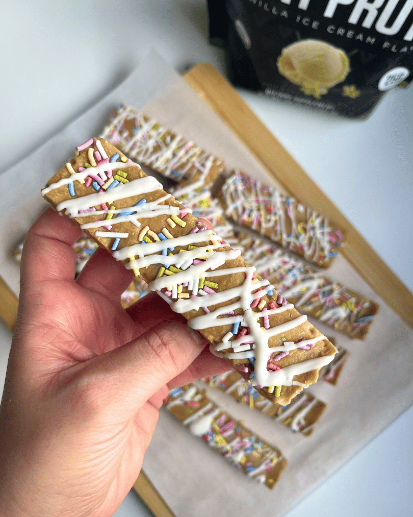 birthday cake protein bar being held by hand close up, and over a wooden cutting board with more bars on it