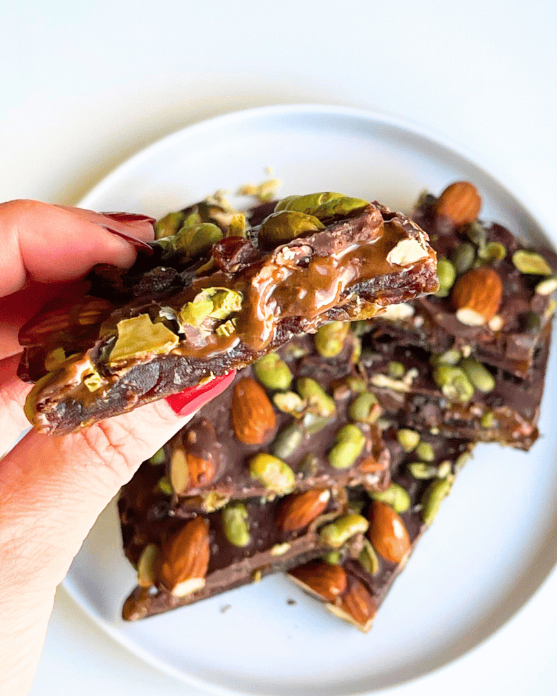 a hand holding slices of protein date bark up close over a plate of them