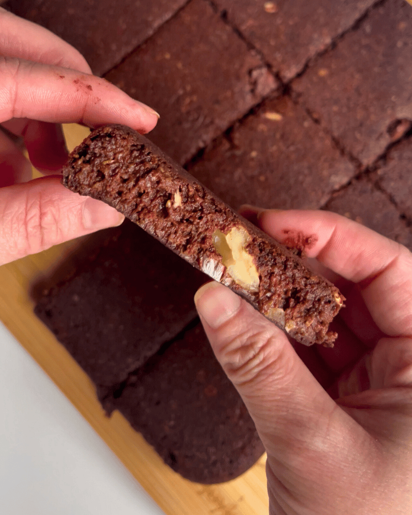 two hands holding a lactation brownie up close over the tray of sliced brownies