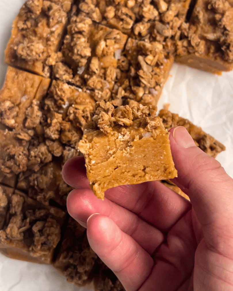 a hand holding a square of maple peanut butter fudge up close over top of the other squares on white parchment paper