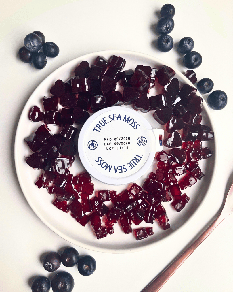 purple vegan gummy bears on a white plate with a bronze spoon laying next to it and blueberries all around