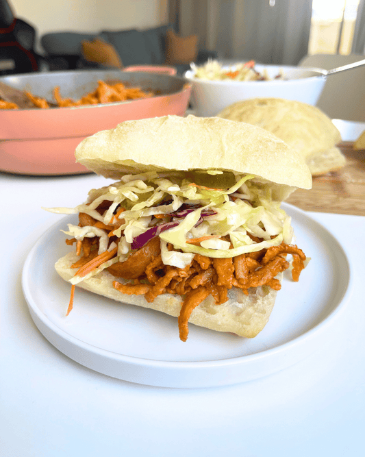 high protein vegan pulled pork sandwich on a white plate with a pot and bowl filled with ingredients in the background
