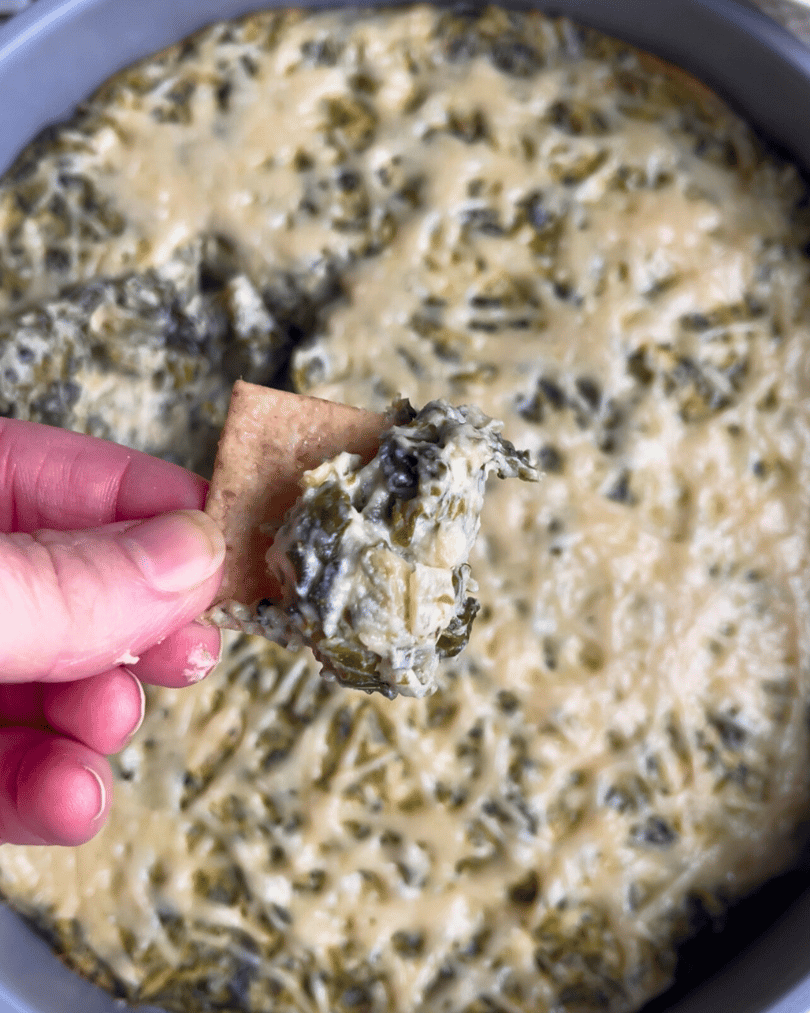 a hand holding a cracker covered in vegan spinach and artichoke dip 