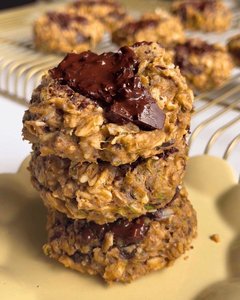 three zucchini protein cookies on top of one another on a small yellow plate with more cookies behind it on a gold cooling rack