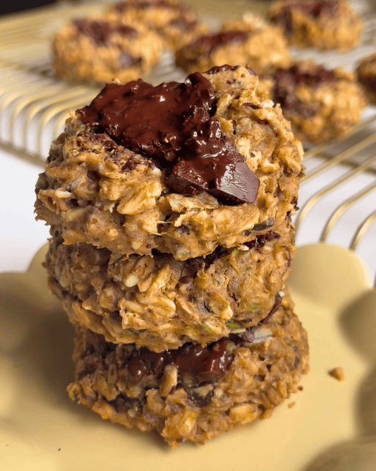 three zucchini protein cookies on top of one another on a small yellow plate with more cookies behind it on a gold cooling rack