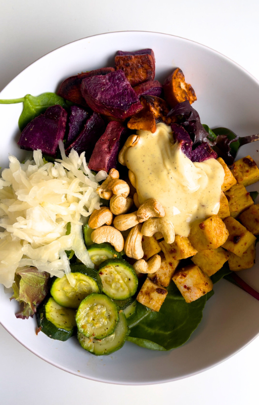 Spinach salad topped with purple yams, baked purple yams, cashews, sauerkraut and baked tofu in a white plate with a white background from the high protein vegan lunch recipes ebook