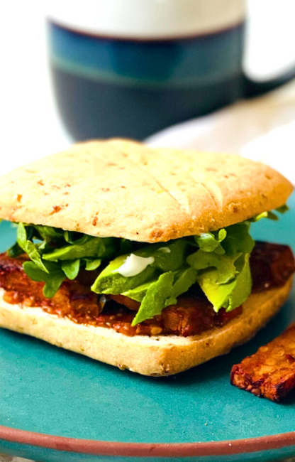 A tempeh sandwich on top of a green plate with a blurry mug in the background